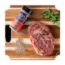 A wooden cutting board holds a seasoned raw steak, a bottle of Rufus Teague Steak Rub by Rufus Teague, a small pile of the spice mix, and a sprig of fresh parsley.
