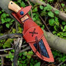 The Texan Knives Custom Handmade 10" Damascus Steel Hunting Knife features an olive wood handle and comes with a brown leather sheath, accented by decorative stitching and a black textured patch, shown on a tree branch with green leaves.