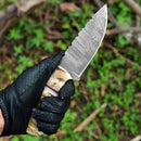 A person wearing a black leather glove holds the Texan Knives Custom Handmade Damascus Steel Skinning Knife with a 4.5-inch sheep horn handle, set against a blurred green outdoor background.