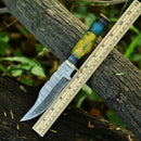 The Texan Knives Damascus Steel Hunting Knife – 10" with Brass Guard features a resin pakkawood handle and comes with a leather sheath, displayed on a log next to a ruler.