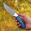 A hand displays the Texan Knives Custom Handmade Damascus Steel Skinning Blade - 4.5" with a blue wooden handle and patterned blade, set against blurred greenery—perfect for outdoor enthusiasts.