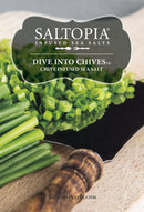 A close-up of chopped fresh chives on a wooden cutting board with a knife and the SALTOPIA Infused Sea Salts label for Saltopia Chive Salt: DIVE INTO CHIVES displayed above the chives.