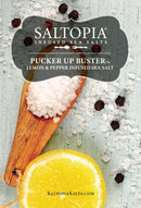 A wooden spoon with coarse sea salt is surrounded by black peppercorns, lemon slices, and scattered salt on a light blue surface. The label reads SALTOPIA Infused Sea Salts Lemon Pepper Salt: PUCKER UP BUSTER.