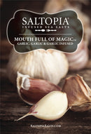 Close-up of garlic bulbs and cloves on wood with text promoting SALTOPIA Infused Sea Salts: Saltopia Garlic Salt—MOUTH FULL OF MAGIC gourmet garlic seasoning. Website SALTOPIASALTS.COM appears at the bottom.