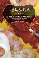 A spoonful of red paprika powder rests on yellow, with onion skins and salt nearby. The label says “SALTOPIA Infused Sea Salts: Saltopia Paprika & Onion Salt: EUREKA SWEET PAPRIKA.”.