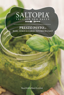 A jar of vibrant green pesto topped with basil sits on wood. The label reads: SALTOPIA Infused Sea Salts — Saltopia Basil, Garlic & Lemon Salt: PRESTO PESTO. Discover this herb-infused seasoning at saltopiasalts.com.