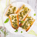 Four slices of toasted bread topped with green herb and garlic spread made using SALTOPIA Infused Sea Salts' Saltopia Italian Herbs Flavor Low Sodium: AMORE are arranged on a white plate, garnished with fresh herbs and set on a wooden surface.
