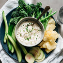A plate with cucumber spears, green beans, broccolini, radish slices, and potato chips surrounds a bowl of creamy dip topped with SALTOPIA Infused Sea Salts' Saltopia Ranch Seasoning Low Sodium: SHOOK UP. A glass of water sits nearby.