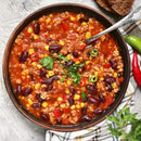 A bowl of chili topped with parsley and SALTOPIA Infused Sea Salts' YACK-ety YACK Southwestern Flavor, featuring kidney beans, corn, ground meat, zesty tomato sauce, red and green chili slices for spice. Bread and spoon nearby on a light surface.