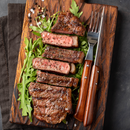A sliced, medium-rare steak rests on a wooden board with arugula and a sprinkle of SALTOPIA Infused Sea Salts’ DIRTY WEDDING Truffle Salt, made with white Italian truffles. Wooden-handled fork and knife, peppercorns, and coarse salt surround the dish.