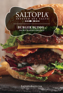Close-up of a gourmet burger with lettuce, cheese, bacon, and two beef patties on a sesame bun. Text overlays promote SALTOPIA Infused Sea Salts’ Saltopia Smoky Blended Salt: BURGER BUDDY as “The Best Burger Seasoning Companion.”.