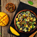 A wooden bowl of quinoa salad with black beans, diced mango, greens, and roasted pumpkin seeds—lightly tossed in SALTOPIA Maple Mustard Seasoning Low Sodium: ANOTHER ONE BITES—sits on a rustic table beside fresh mango and pumpkin seeds.