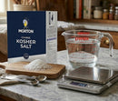 A box of Morton coarse kosher salt, a pile of salt on a wooden board, measuring spoons, a Pyrex cup filled with water, and a digital scale on a marble counter—the perfect essentials for mastering brisket injection or meat injecting techniques straight from SpitJack’s “The Ultimate Guide to Meat Injecting” PDF eBook.