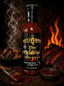 A bottle of Scott’s Smoked Q The Original Sweet Garlic BBQ Sauce is displayed alongside smoked ribs, a bowl of rich BBQ sauce, a brush, wood chips, and a crackling fire in the background. Swirling smoke fills the air, creating a rustic and smoky ambiance.