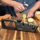 A NULU Inc. Adjustable Knife Sharpener rests on a wooden counter, its clearly labeled slots for angle, coarse, fine, and scissors making it a versatile tool for any kitchen. In the softly blurred background, someone slices cucumbers and carrots.