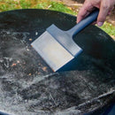 A hand uses a wide stainless steel Fox Run Brands Griddle Scraper to clean a dark, seasoned cooking surface.