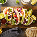 Three soft tacos filled with shredded meat, avocado, tomato, corn, red cabbage, cilantro, and cheese on a wooden board—seasoned with SALTOPIA Infused Sea Salts' HOLY MOLY Taco Seasoning for authentic flavor. Lime wedges and sauce are served alongside.
