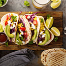 Three tacos with shredded meat, avocado, tomato, corn, purple cabbage, cilantro, and cheese on a wooden board are finished with SALTOPIA Infused Sea Salts’ Saltopia Lime Garlic Salt. Lime wedges, sauce, tortillas, and a drink are nearby.