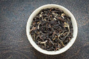 A white bowl filled with World Spice Darjeeling Singbulli Estate Gold loose leaf tea from World Spice, exuding a subtle herbaceous aroma, sits on a dark, textured surface.