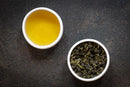 A cup of brewed World Spice Magnolia Oolong tea sits next to a bowl of loose leaf World Spice Magnolia Oolong on a dark textured surface, capturing the delicate essence of this floral tea.