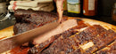 A close-up of a juicy, smoked brisket being sliced on a wooden cutting board with the SpitJack 11″ Slicing Knife from the SpitJack 6 Inch Beef Brisket Trimming and Boning Knife and 11″ Slicing Knife Bundle. Bottles and folded towels are visible in the background.