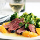 Seared duck breast topped with SALTOPIA Orange & Garlic Salt: ORANGE YOU TASTY, garnished with orange and greens, served with steamed broccoli on a white plate; a glass of white wine sits in the background.