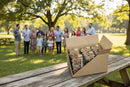 A bulk case of Texas Pecan Cakes— the Texas Pecan Cake 30-Pack— rests on a picnic table in a sunlit park, surrounded by a group of adults and children gathered together in the background.