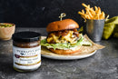 A jar of Terrapin Ridge Farms Balsamic Onion Jam sits in front, ideal for cheese boards. In the background, a cheeseburger with lettuce and a side of fries in a metal cup are served on a plate.