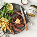 A plate with sliced steak topped with butter, fries, fresh greens, and a lemon wedge sits beside Terrapin Ridge Farms Smokey Maple Bacon Mustard—a great addition to charcuterie boards—plus cutlery and a salt bowl on a marble surface.