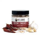 A clear jar of B.T. Leigh’s Aleppo & Garlic Finishing Salt sits on a white surface, surrounded by three dried chili peppers and a whole head of garlic with several loose cloves.