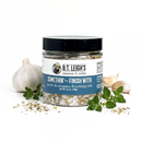 A jar of B.T. Leigh’s Garlic & Oregano Finishing Salt surrounded by a head of garlic, cloves, and fresh herbs.