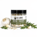 A clear jar of B.T. Leigh’s Garlic & Rosemary Finishing Salt surrounded by a whole garlic bulb, loose cloves, and sprigs of fresh rosemary.