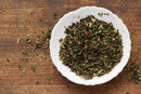 A white bowl filled with World Spice Spearmint dried green leaves from World Spice sits on a rustic wooden surface—perfect for brewing mint tea or enhancing spice blends—with a few leaves scattered around the bowl.