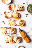 Four toasted baguette slices with poached eggs, tomatoes, and microgreens are drizzled with Terrapin Ridge Farms Spicy Chipotle Garnishing Squeeze 14 oz for smoky chipotle flavor, arranged on white marble with the Terrapin Ridge Farms bottle nearby.