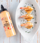 A bottle of Terrapin Ridge Farms Sriracha Horseradish Garnishing Squeeze Sauce sits next to a white dish with three shrimp, each topped with the creamy sauce and chopped herbs, displayed on a light wooden surface.