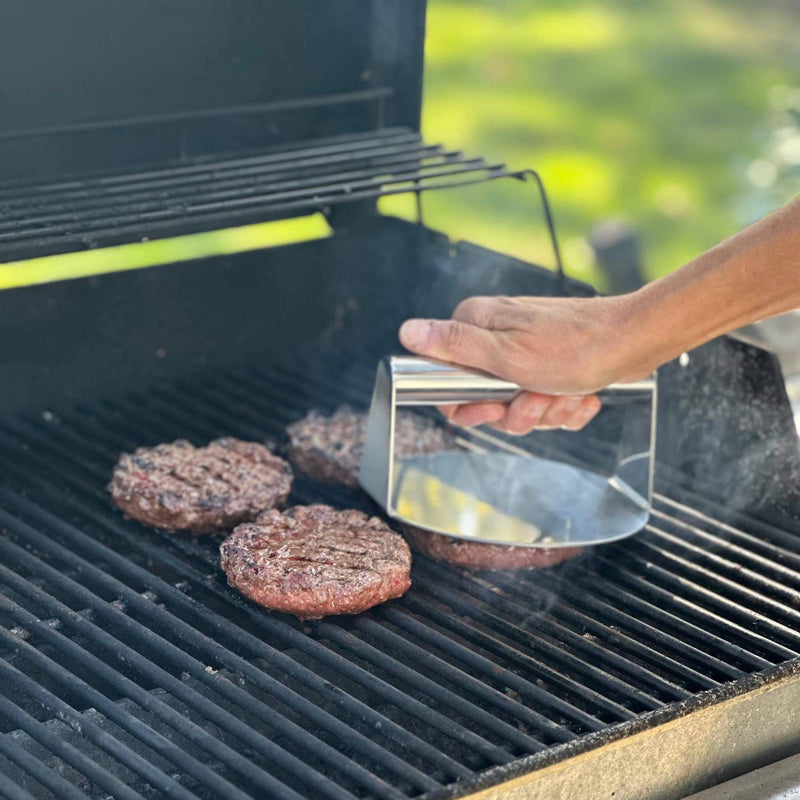 stainless steel burger smasher grill press 5.7 x 6 inches with ergonomic handle shown with burgers on charcoal grill 