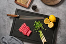 A Steelport Knife Co. 10" Slicing Knife rests on a cutting board alongside sliced raw tuna, chopped green onions, whole green onions, and two halved lemons. Small dishes of sauces and seasonings are arranged nearby on a gray tabletop.