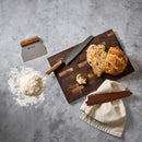 The Steelport Knife Co. 3-Piece Baker Set by Steelport Knife Co.—featuring a bread knife, dough scraper, and other premium tools—rests on a wooden board alongside a loaf of bread, flour pile, and towel on a gray countertop.