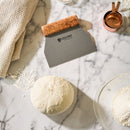 A ball of dough rests on a floured marble surface alongside the Steelport Knife Co. 3-Piece Baker Set—a collection of handcrafted kitchen tools from Steelport Knife Co.—with a kitchen towel, copper measuring cup, and a glass bowl of flour nearby.