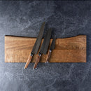Three Steelport Knife Co. 3-Piece Essential Set kitchen knives with wooden handles rest on a rectangular wooden cutting board, which is set atop a dark textured surface.