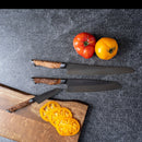 Two Steelport Knife Co. 3-Piece Essential Set kitchen knives with wooden handles rest on a gray countertop near a red and a yellow tomato, with one Steelport knife placed beside sliced yellow tomato on a wooden cutting board.