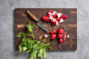 A wooden cutting board with the Steelport Knife Co. 4" Paring Knife, whole and chopped radishes, and radish greens on a gray countertop.