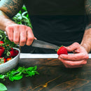 A person holds a strawberry and uses the Steelport Knife Co. 4" Paring Knife to remove its stem, while a bowl of strawberries and fresh mint leaves rest on the wooden table nearby.