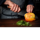 A person wearing a striped apron slices a large yellow tomato with the Steelport Knife Co. 8" Chef Knife, celebrated for its exceptional blade edge retention from traditional knife forging, on a wooden surface. Chopped greens are visible in the foreground.