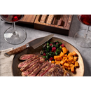 A plate with sliced steak, diced sweet potatoes, and sautéed greens sits beside a knife from the Steelport Knife Co. 8-Piece Steak Knife Set, two glasses of red wine, and a wooden box on a gray table.