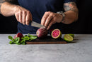 A person slices a prickly pear on a mini cutting board using a paring knife from the Steelport Knife Co. Bar Set, while mint leaves, a halved lime, and another whole prickly pear rest on the table. The individual, who sports tattoos, wears a watch and a dark shirt.