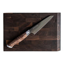 A chef’s knife with a wooden handle rests diagonally on a dark wooden cutting board alongside a sleek paring knife. The board, reminiscent of the craftsmanship found in the Steelport Knife Co. Bar Set from Steelport Knife Co., features a rich, checkered pattern and rounded edges.