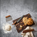 A bread loaf and slices are displayed on a cutting board alongside a bread knife with an ergonomic handle, the Steelport Knife Co. Bench Scraper featuring a stainless steel blade resting on flour, and a cloth-wrapped knife sheath, all arranged on a gray surface.
