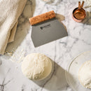 A ball of dough rests on a floured marble surface alongside the Steelport Knife Co. Bench Scraper by Steelport Knife Co., featuring an ergonomic handle and stainless steel blade, as well as a measuring cup, a glass bowl with flour, and a textured kitchen towel nearby.