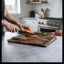 Using a Steelport Knife Co. Signature Knife & Board Set, a person slices an orange on a wooden cutting board in a modern kitchen. Fresh herbs and kitchen utensils from Steelport Knife Co. are visible on the counter near the stove.